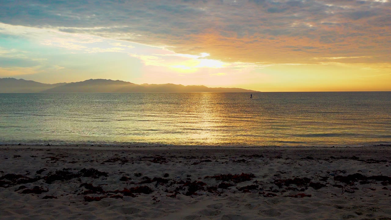A warm sunset spreads under a cloud layer over the sea at El Sargento, Mexico, with silhouetted landforms left of frame, soft ripples, and no visible people.