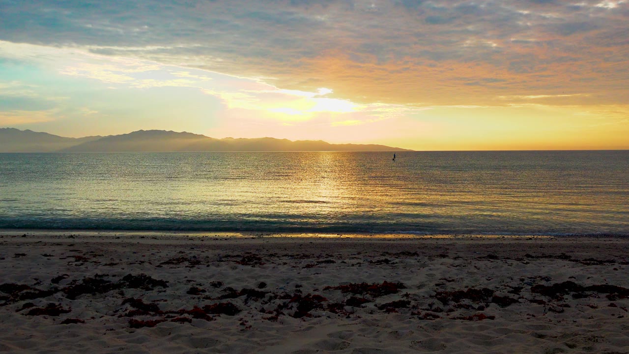 El Sargento La Ventana, Mexico sunset over calm water, with seaweed on the dark sand foreground, distant mountain silhouettes, gold reflection on the sea, and no visible people.