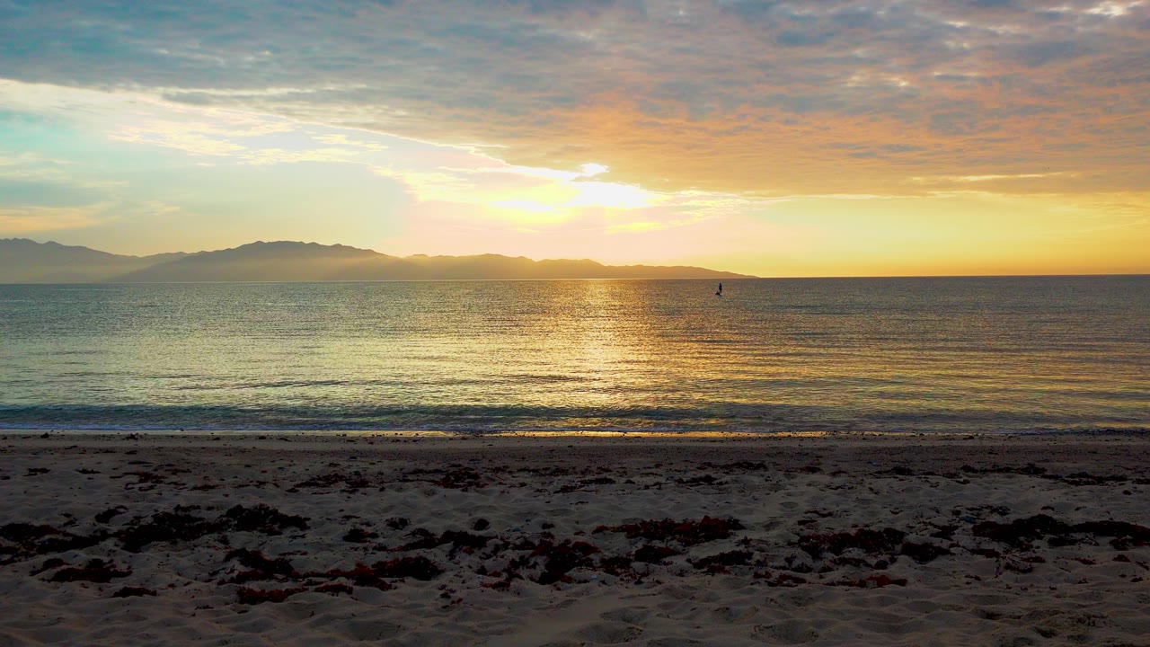 El Sargento La Ventana, Mexico sunset frame with shadowed beach sand, a narrow wave line, calm sea, distant mountains, warm clouds, and no visible people.