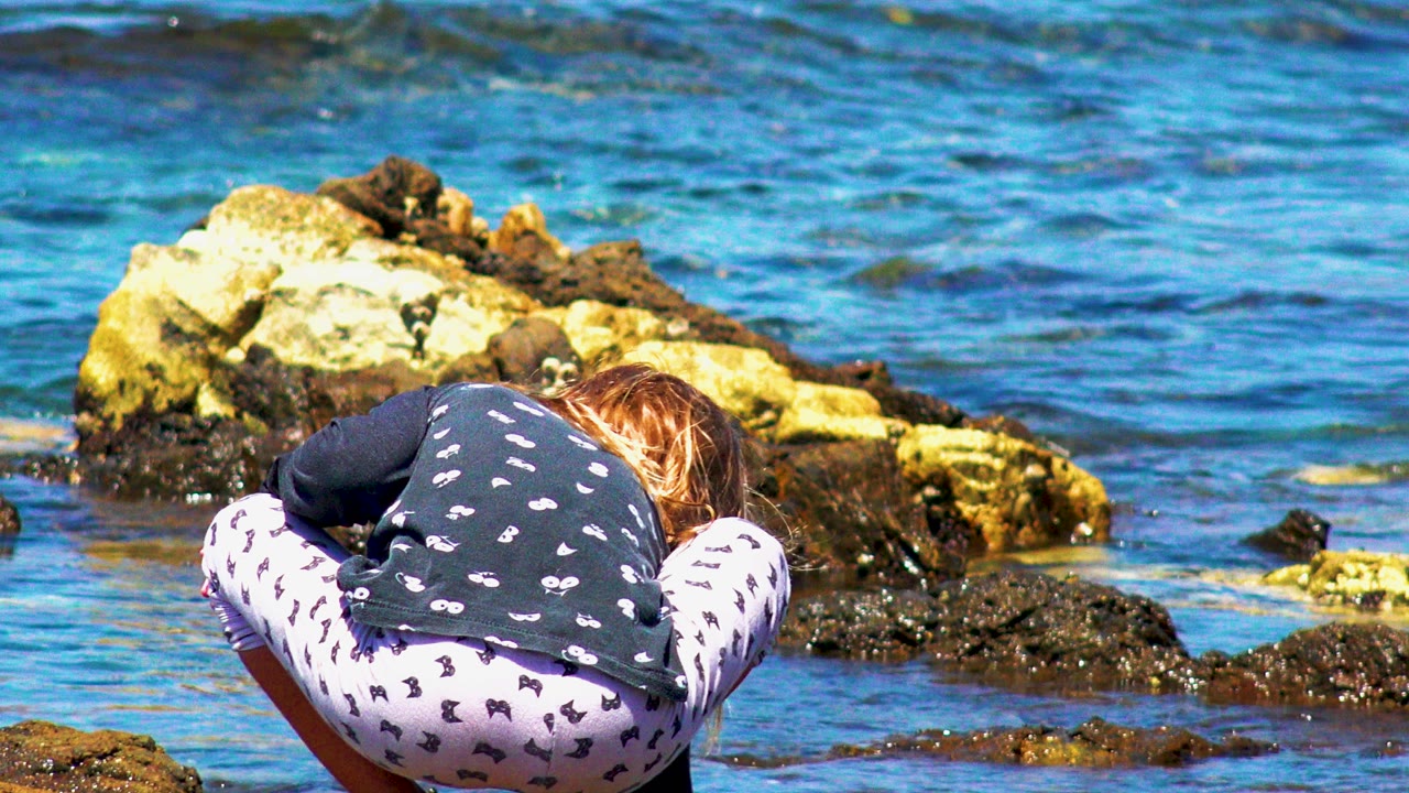 At 0:00, a child crouches with her back to camera near jagged shoreline rocks and bright blue water in El Sargento La Ventana, Mexico.