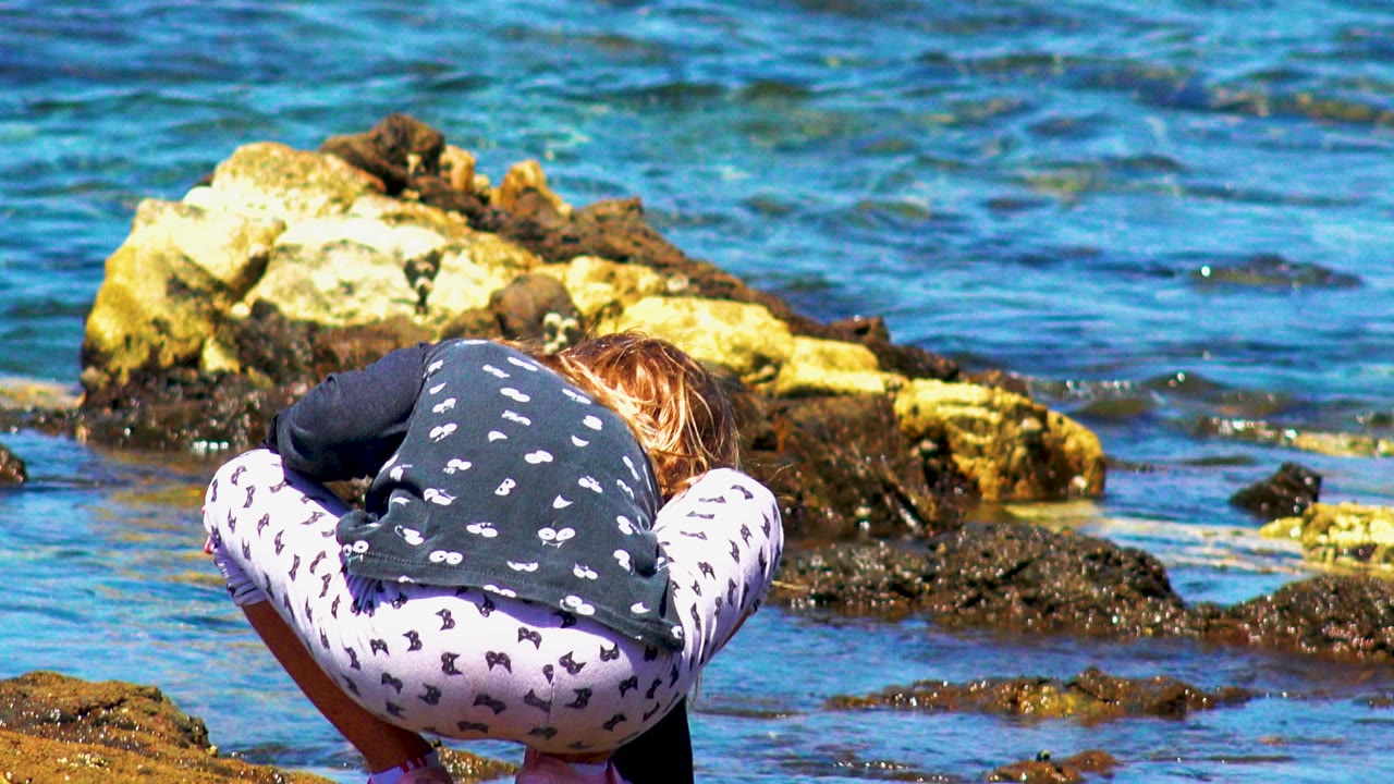 At 0:02, the crouching child is centered lower in frame, with pale rocks behind her and blue rippled water spread across the background in El Sargento La Ventana, Mexico.