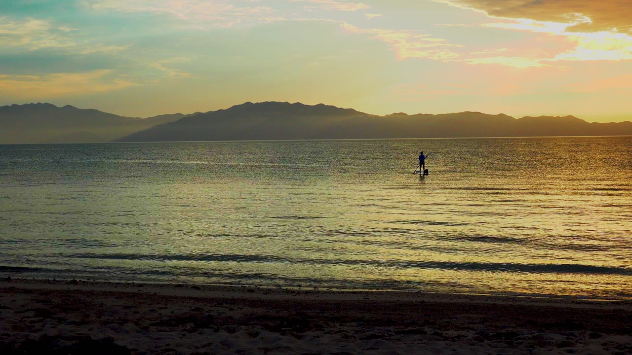 At 0:00 in El Sargento La Ventana, Mexico, a lone paddle boarder stands near the center of calm water as orange sunset light spreads across the ocean and mountains sit on the horizon.