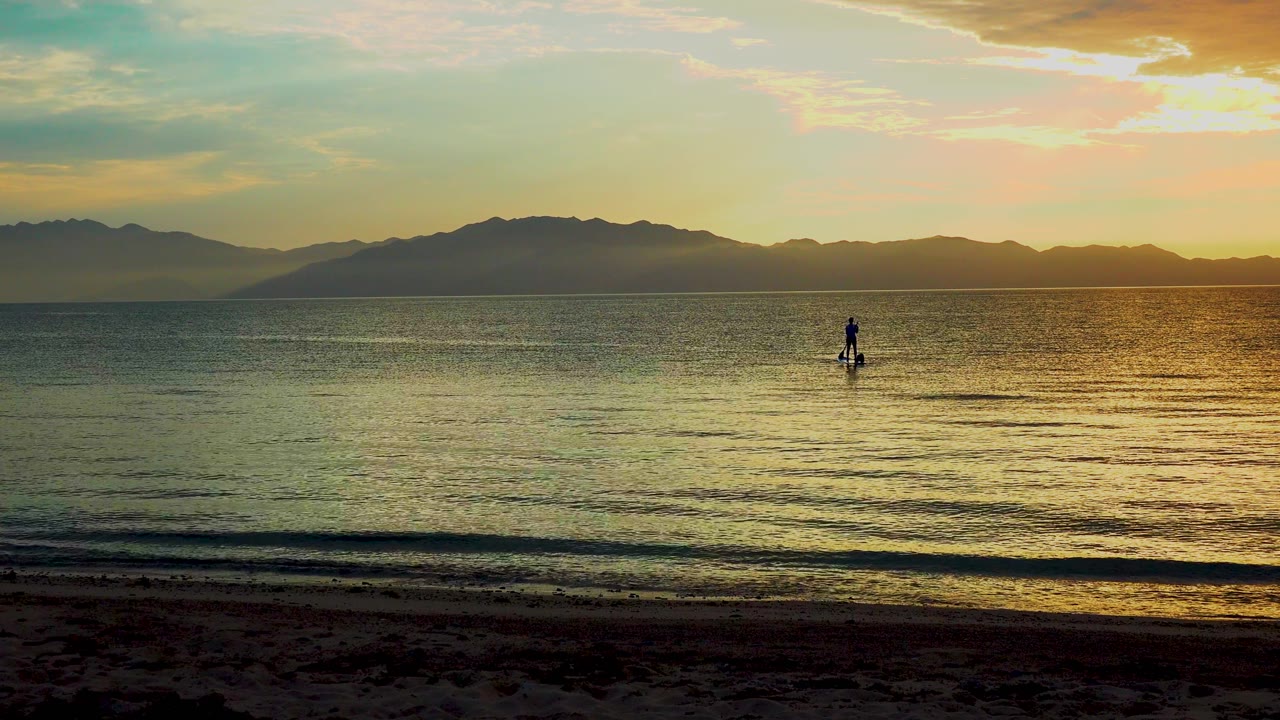 At 0:02 in El Sargento La Ventana, Mexico, the paddle boarder appears slightly right of center on golden water, with low waves near the dark beach and mountains under a glowing sky.
