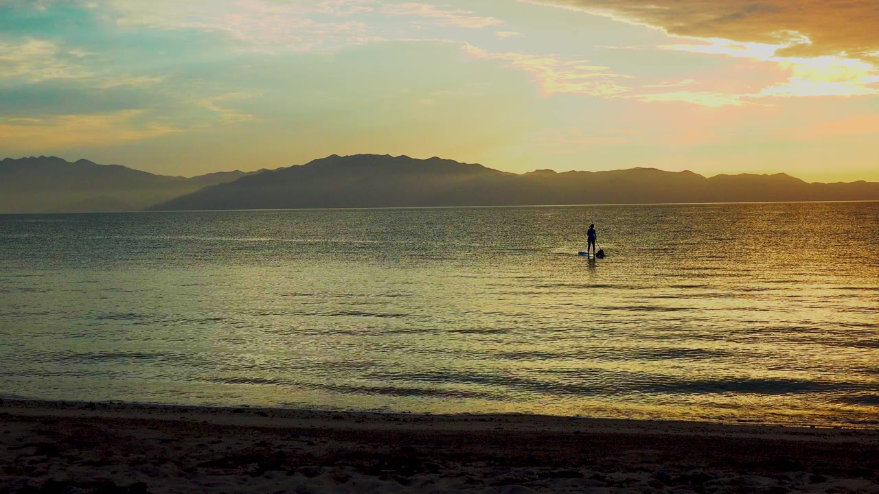 A distant paddleboarder stands on calm rippled water in El Sargento La Ventana, Mexico, with a dark beach strip below and warm sky over the mountain horizon.