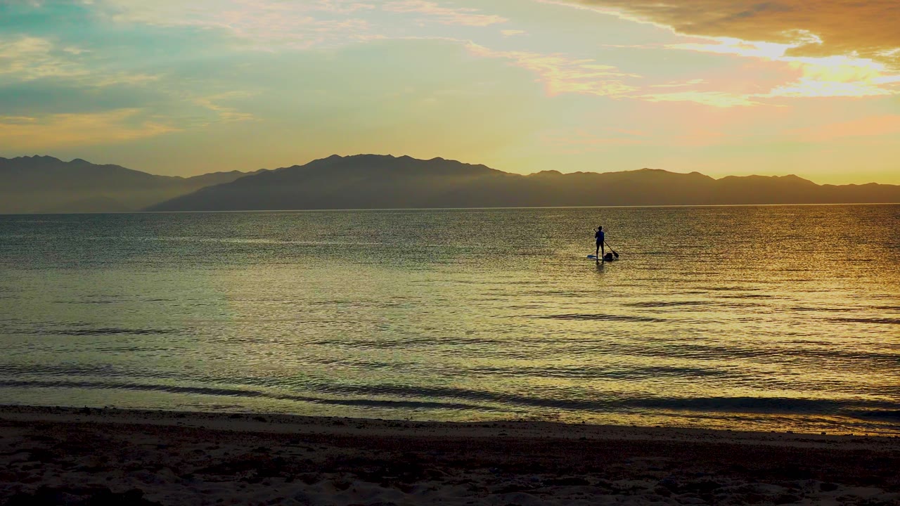 A small paddleboarder moves across shallow wave lines near shore, with the brightest gold water on the right and mountains spanning the background in El Sargento La Ventana, Mexico.