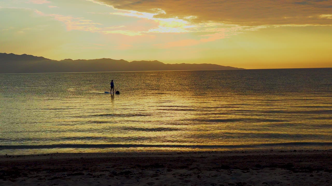 A paddle boarder stands far from camera in shallow water at El Sargento La Ventana, Mexico, with golden sunset reflection across the sea and mountains stretching along the horizon.