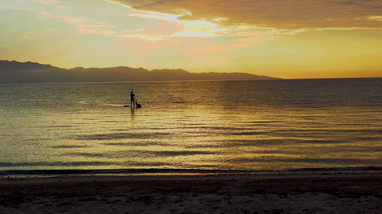 A small paddle boarder silhouette stands on reflective water in El Sargento La Ventana, Mexico, with the brightest sunset glow above the horizon and darker mountains to the left.