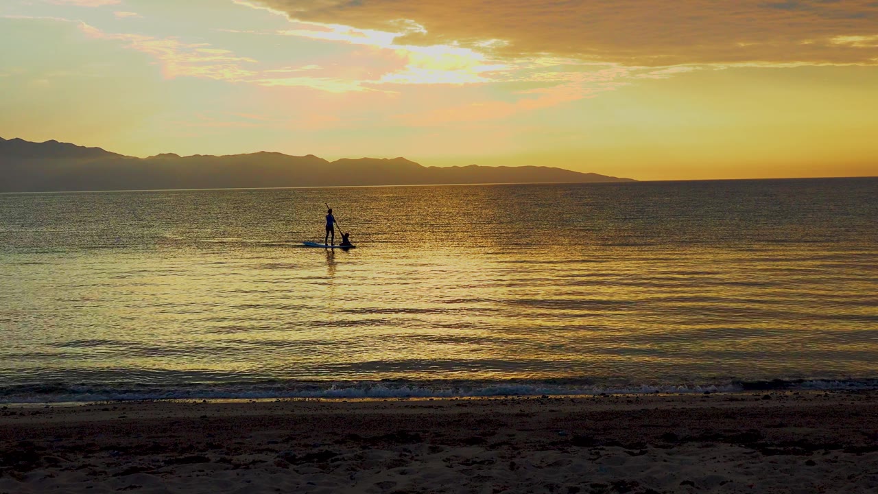 A small silhouetted paddleboarder appears near center frame in El Sargento La Ventana, Mexico, with low waves, orange sky, and dark mountains in the distance.