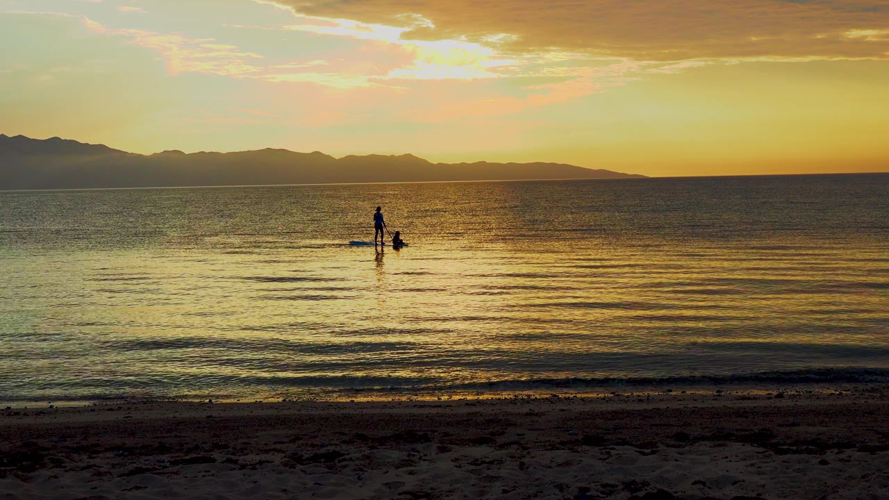A lone silhouetted figure remains beside the kayak in shallow water at El Sargento La Ventana, Mexico, under a bright warm cloud band.
