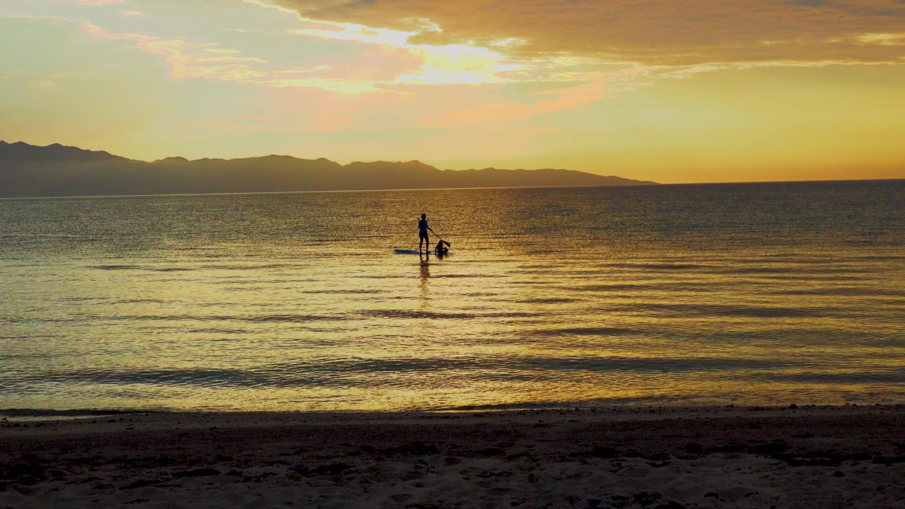 At 0:00, a distant person paddles on calm water in El Sargento La Ventana, Mexico, with a dark beach foreground and low mountains under golden sky.
