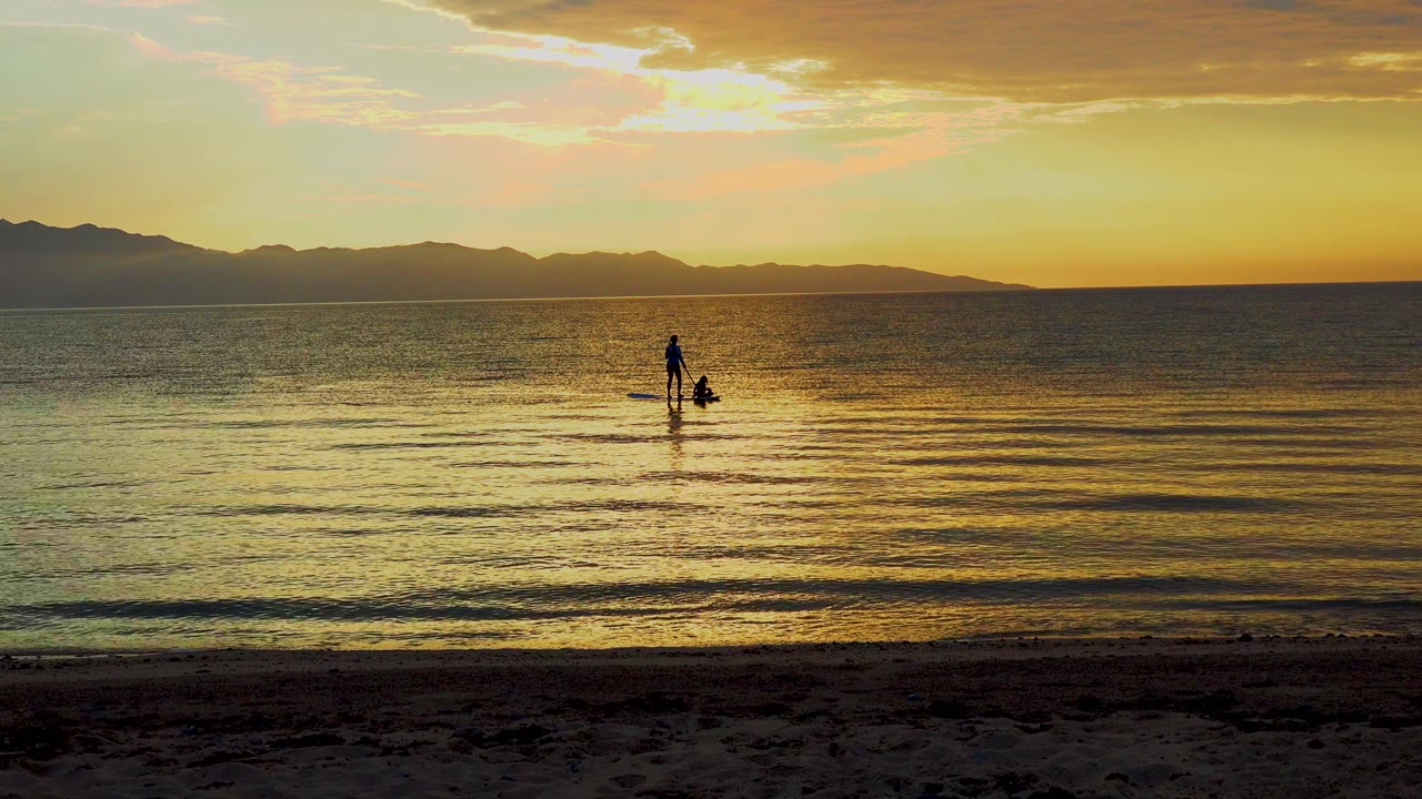 At 0:02, the paddler appears slightly right of center on the gold-lit water, with low ripples, a dark sandy foreground, and a continuous mountain ridge in back.