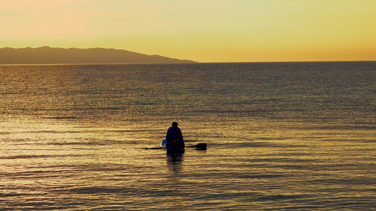 Small boat with one person on golden water under a distant coastline.