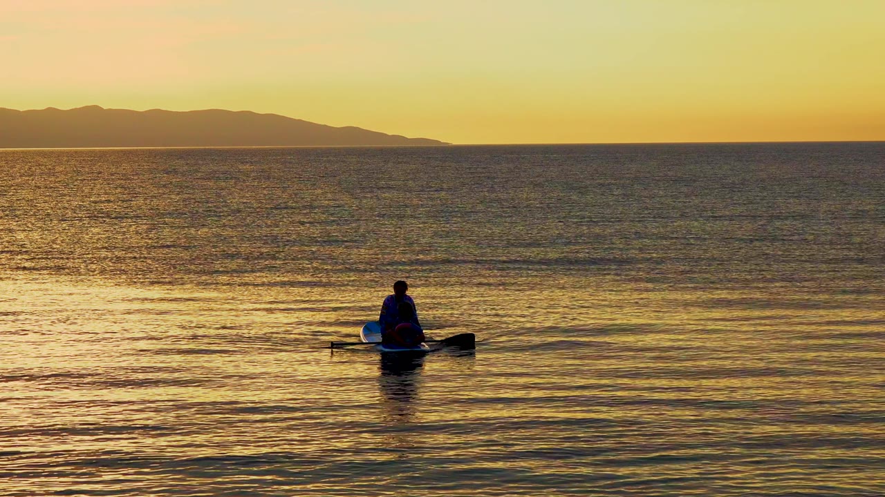 A seated person on a small boat appears slightly right of center on golden ocean water, with a long dark land silhouette under a yellow sky in El Sargento La Ventana, Mexico.