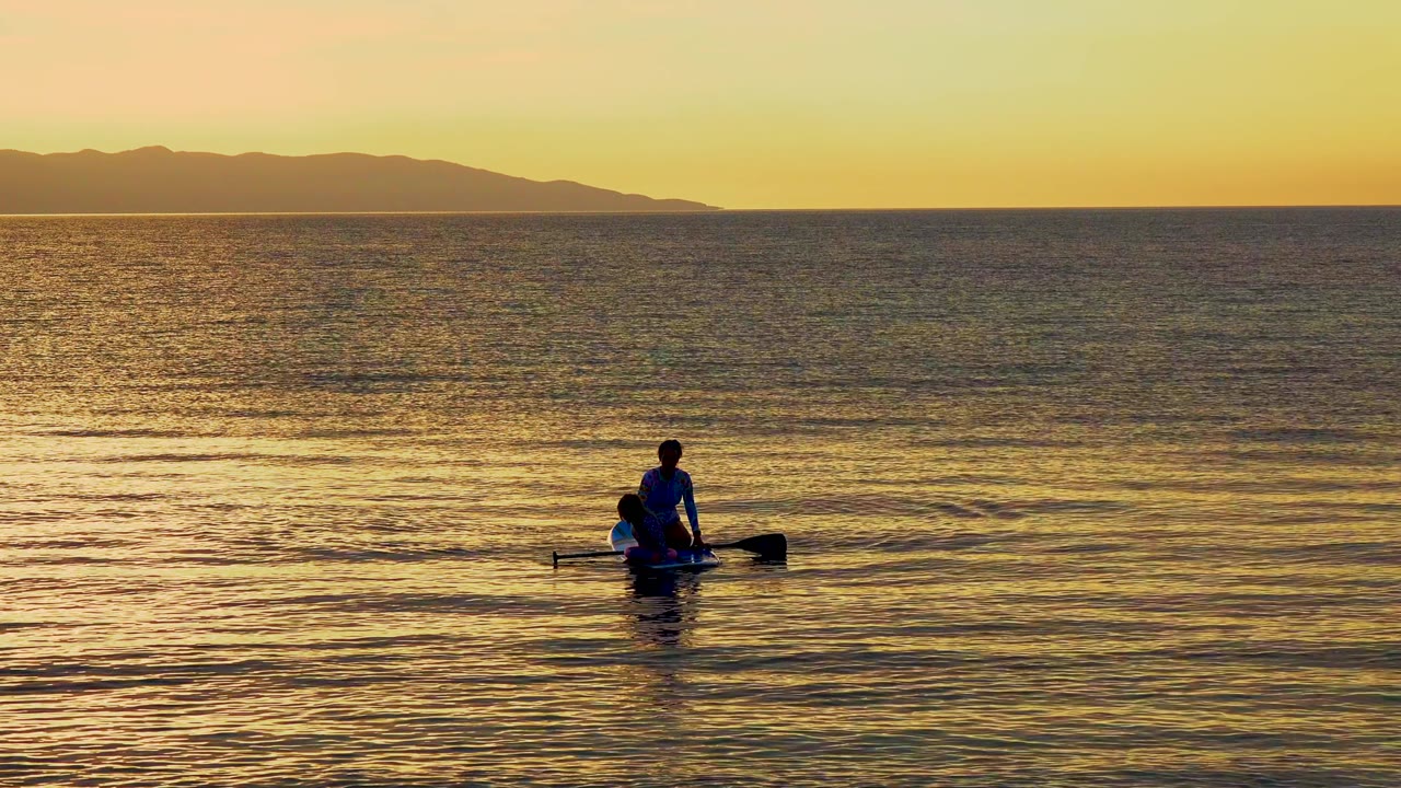 A backlit person in a small boat rows across warm rippled water in El Sargento La Ventana, Mexico, with dark hills spanning the upper background.