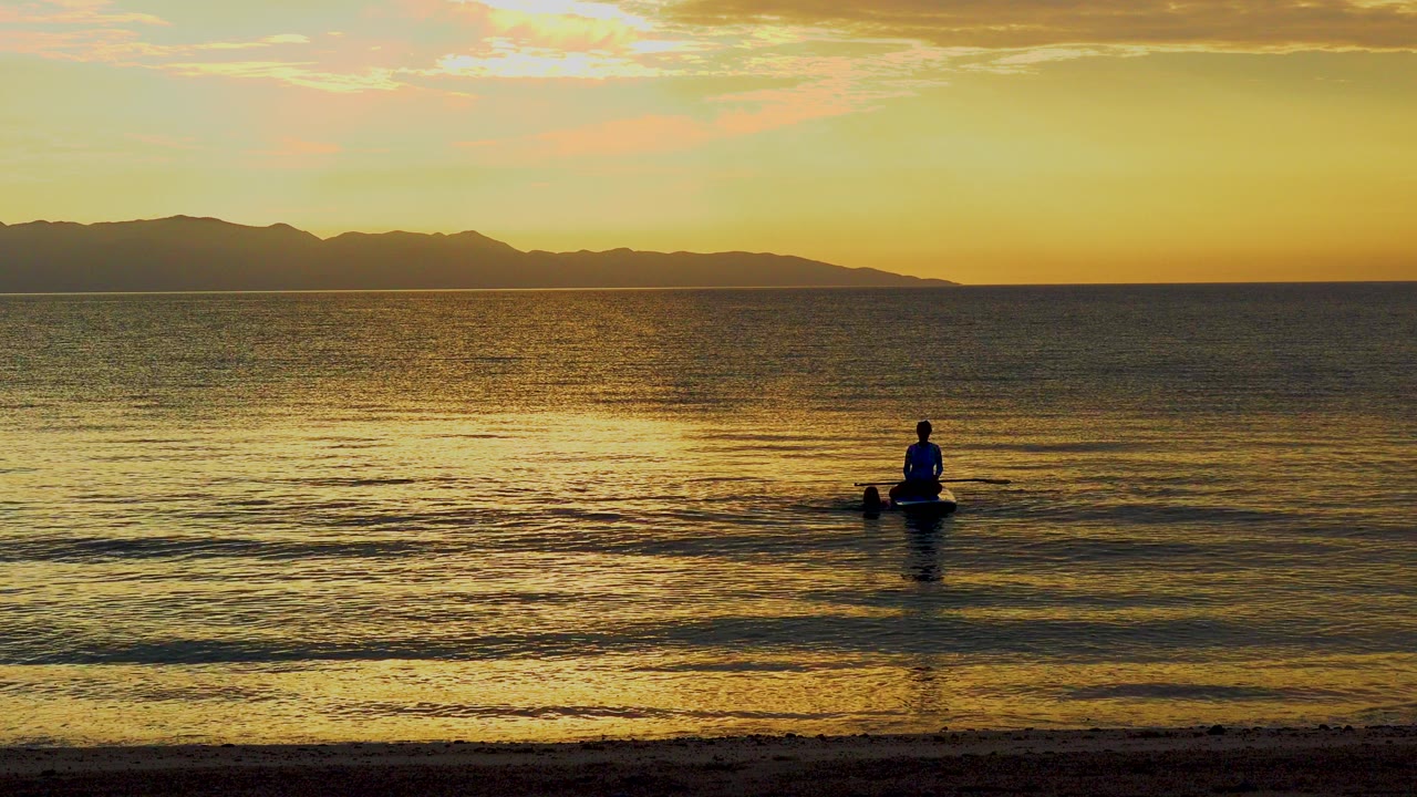 A distant paddler floats near shore on calm sunset water in El Sargento La Ventana, Mexico, with a dark mountain ridge across the horizon.