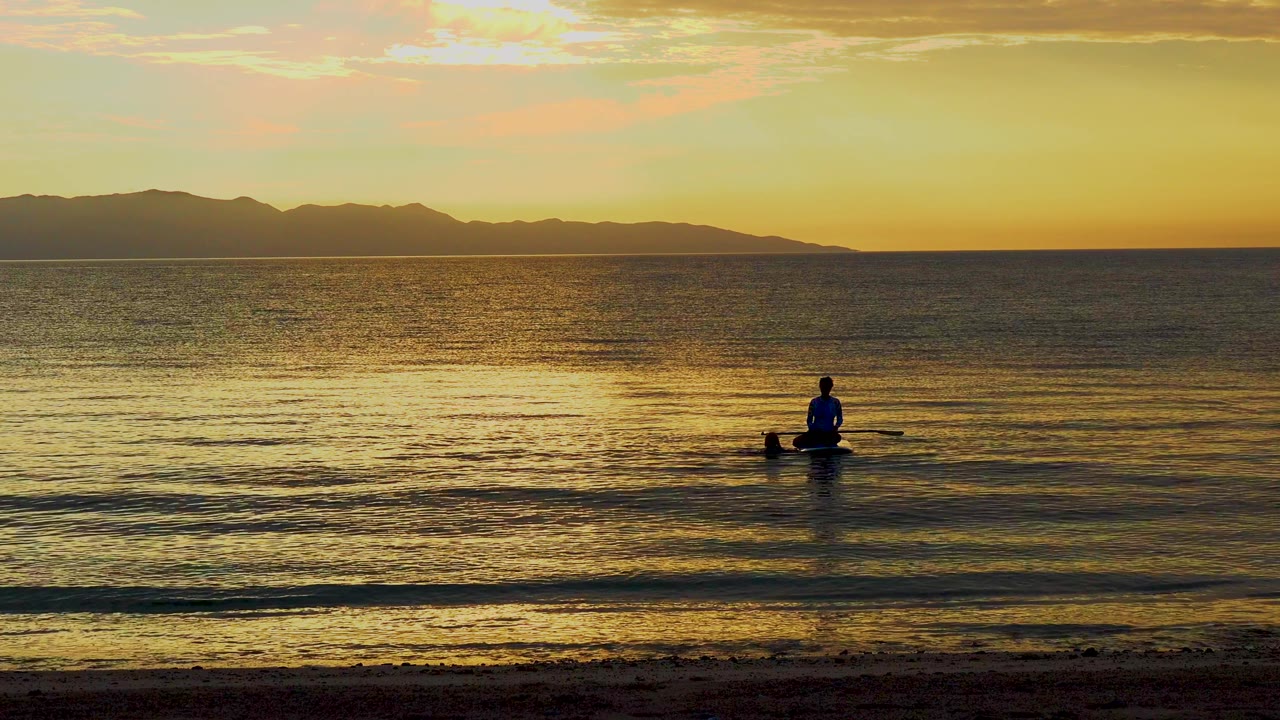 A small paddler sits slightly right of center on the calm water, with a thin beach line below and the Mexico mountain horizon dark against the orange sky.
