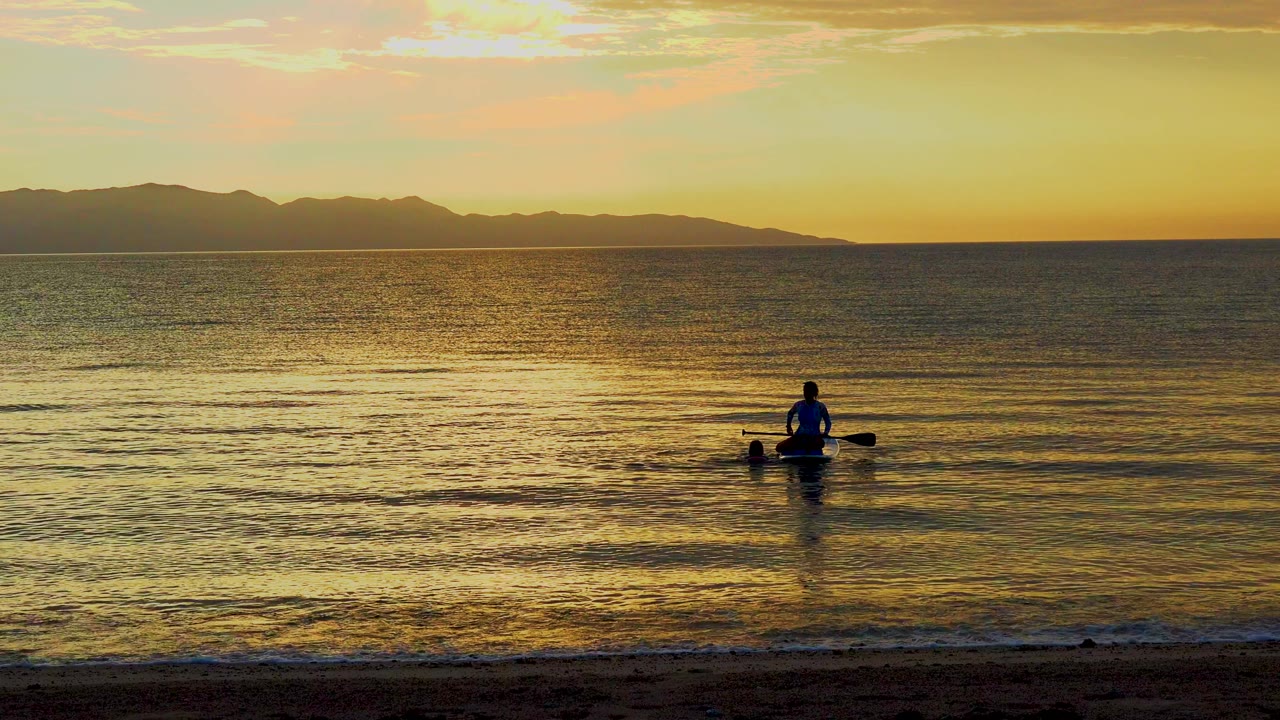 A distant silhouetted paddler sits near the center of reflective water in El Sargento La Ventana, Mexico, with a dark beach foreground and mountain horizon behind.