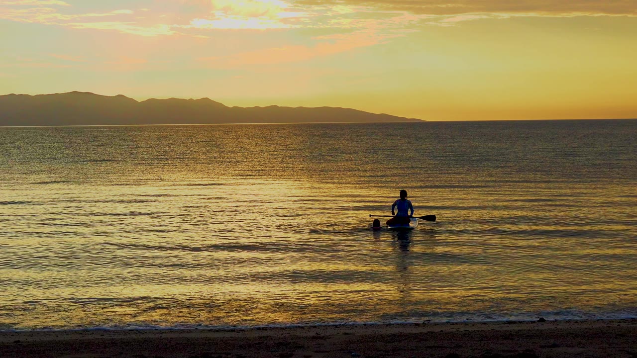 The paddler remains small against the wide golden water in El Sargento La Ventana, Mexico, with soft ripples spreading toward the beach and mountains across the background.
