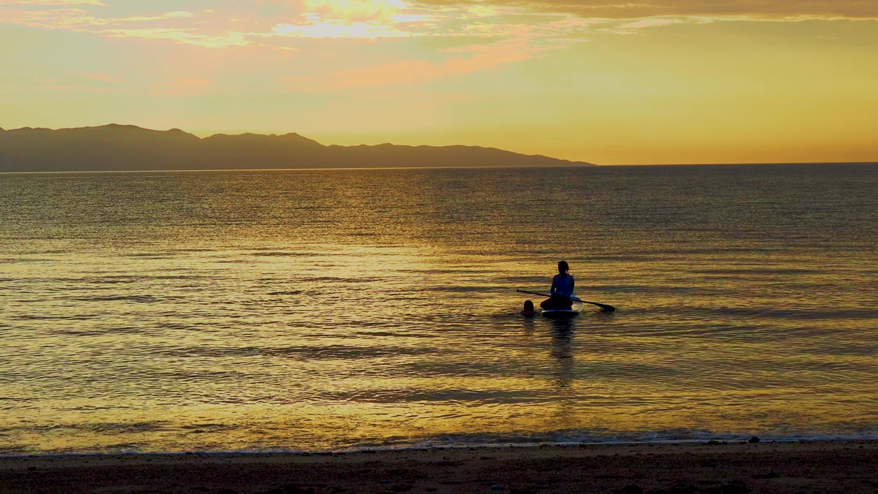 A distant paddler shifts farther right in the frame while golden light streaks across the water and the mountain ridge stays dark along the horizon in El Sargento La Ventana, Mexico.
