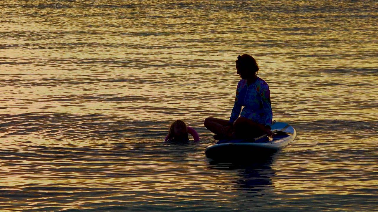 At 0:00 in El Sargento La Ventana, Mexico, a silhouetted person sits cross-legged on a paddleboard while another person floats nearby in golden rippled water.
