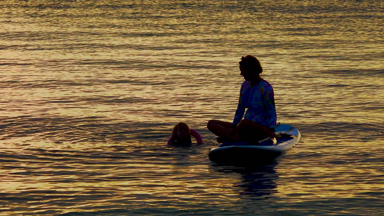 Seated paddleboarder in silhouette beside a floating person on warm sunset water.