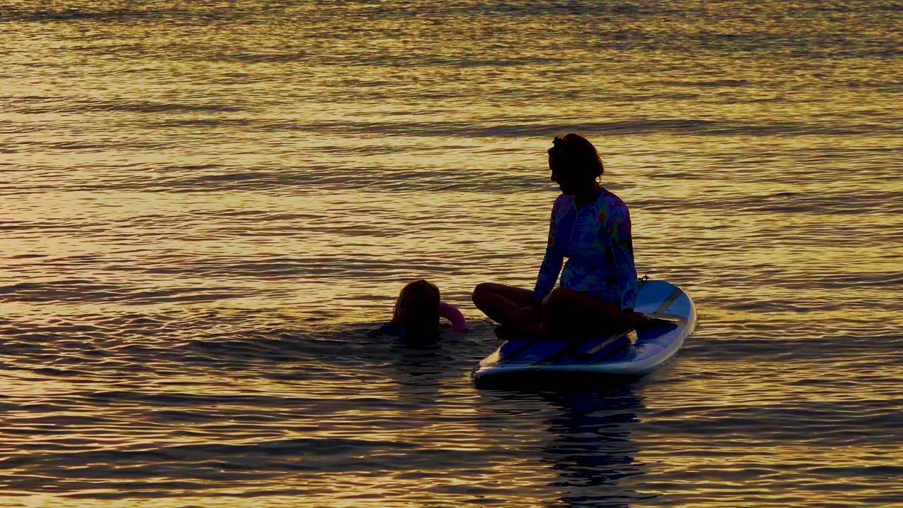 At 0:02 in El Sargento La Ventana, Mexico, the paddleboarder sits to the right of the floating person as low waves break the orange-gold reflection pattern.