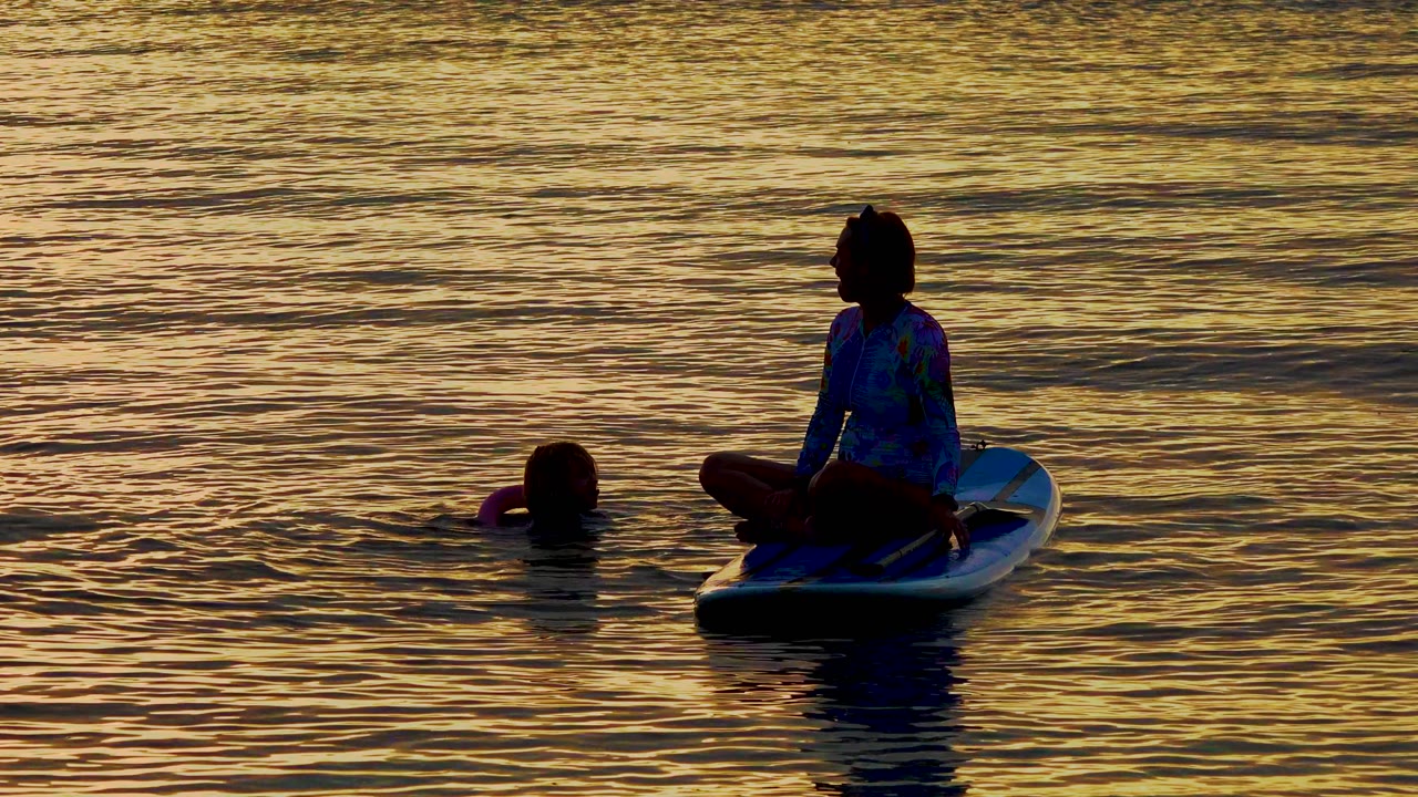 At 0:00 in El Sargento La Ventana, Mexico, a silhouetted woman sits cross-legged on a small board while a child floats nearby in warm rippled water.