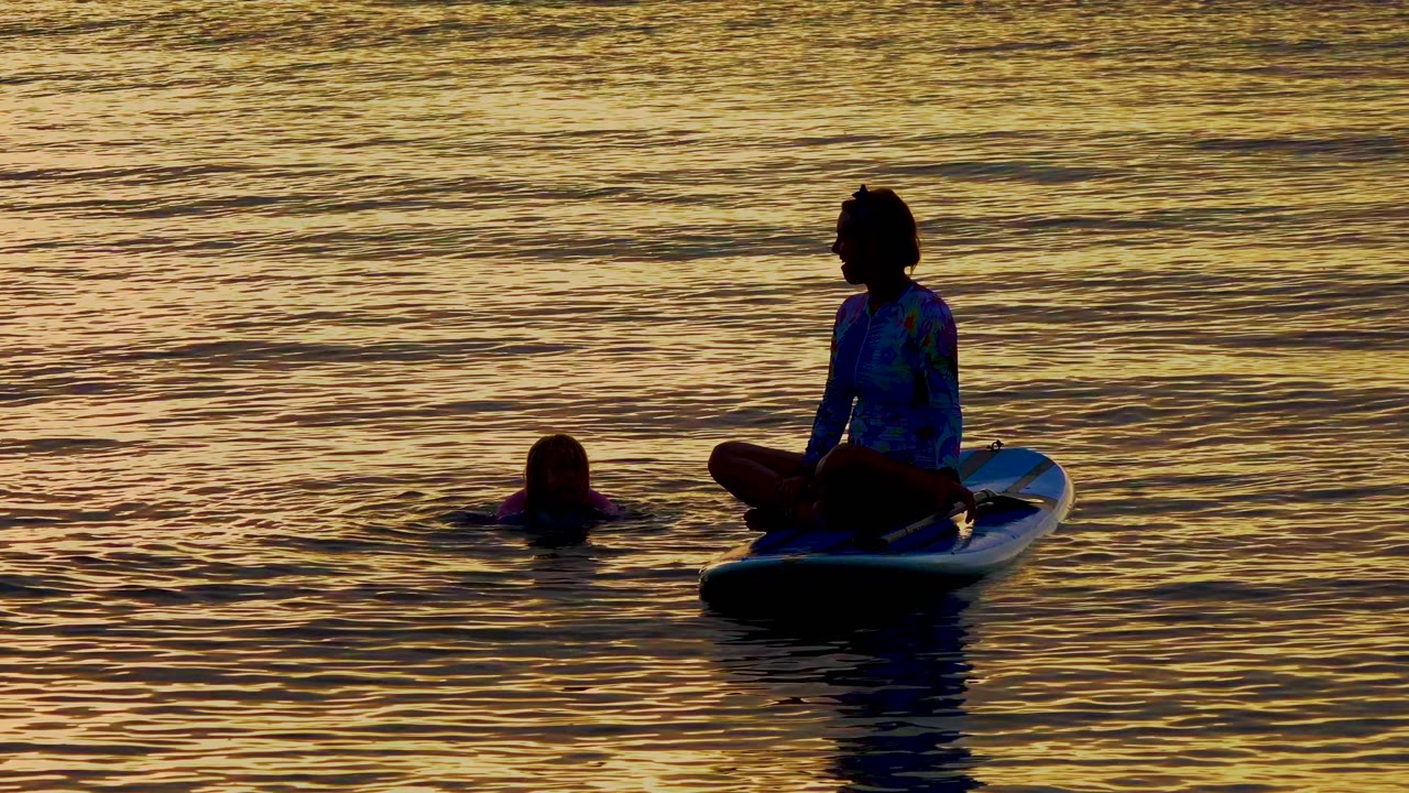 At 0:02 in El Sargento La Ventana, Mexico, the woman sits in profile on the board while the child's head and flotation device remain visible in the surrounding dusk water.