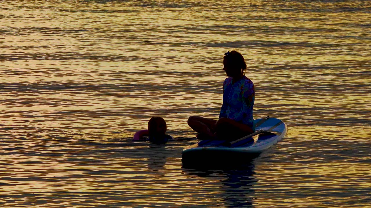At El Sargento La Ventana, Mexico, a silhouetted person sits cross-legged on a white board while another person floats to the left in gold-reflecting ocean water.