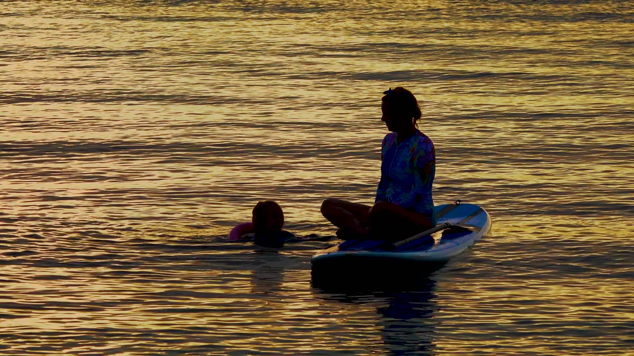 At 0:01, the seated person remains centered on the board while the swimmer stays close on the left and narrow gold ripples cross the water behind them.