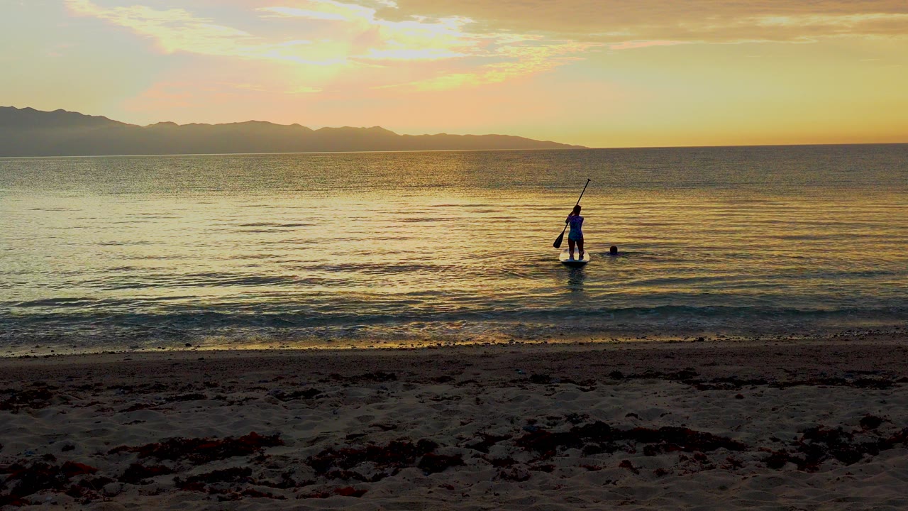 A distant paddleboarder stands near the shoreline in El Sargento La Ventana, Mexico, with orange sunset light on calm water and dark beach foreground.
