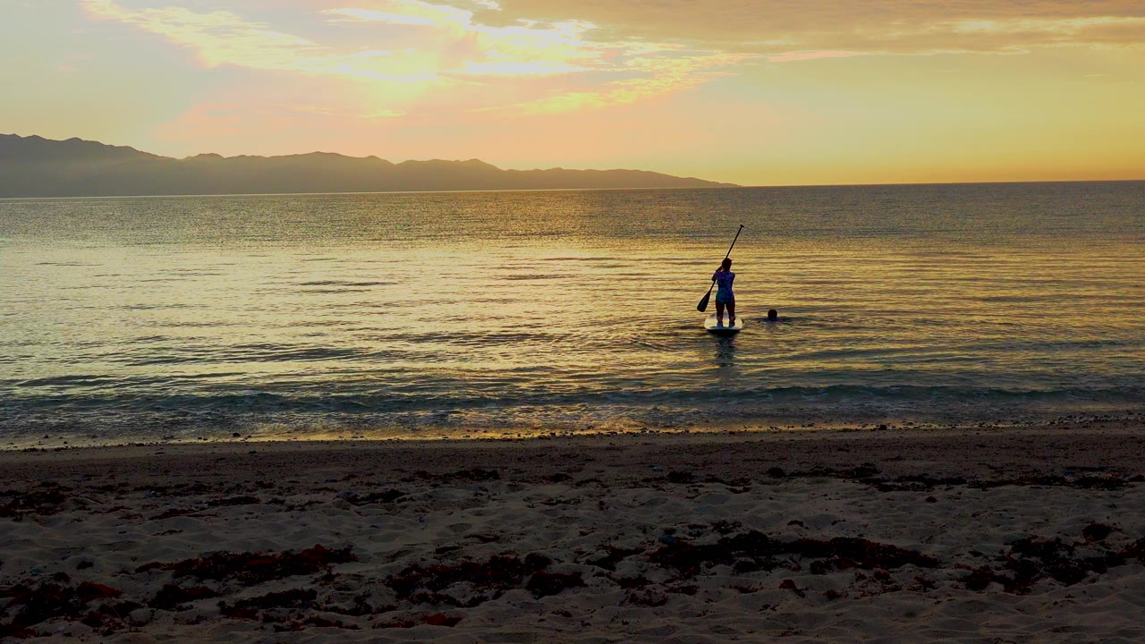 The paddleboarder remains near the center-right of the frame in El Sargento La Ventana, Mexico, with golden water ripples and a dark strip of beach below.
