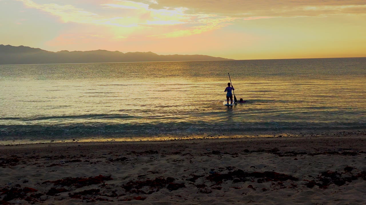 A small standing paddleboarder appears farther right in the composition at El Sargento La Ventana, Mexico, with low waves and a wide orange-blue horizon behind them.