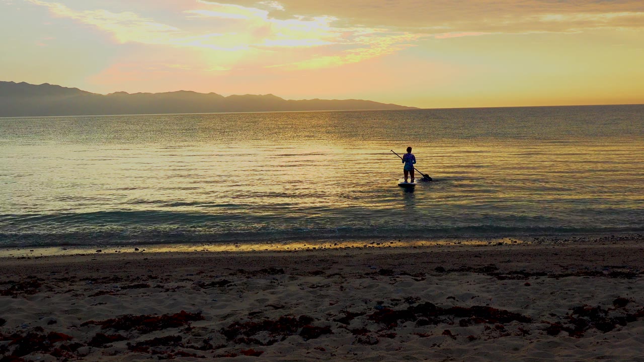Paddleboarder silhouetted in shallow water under sunset sky.