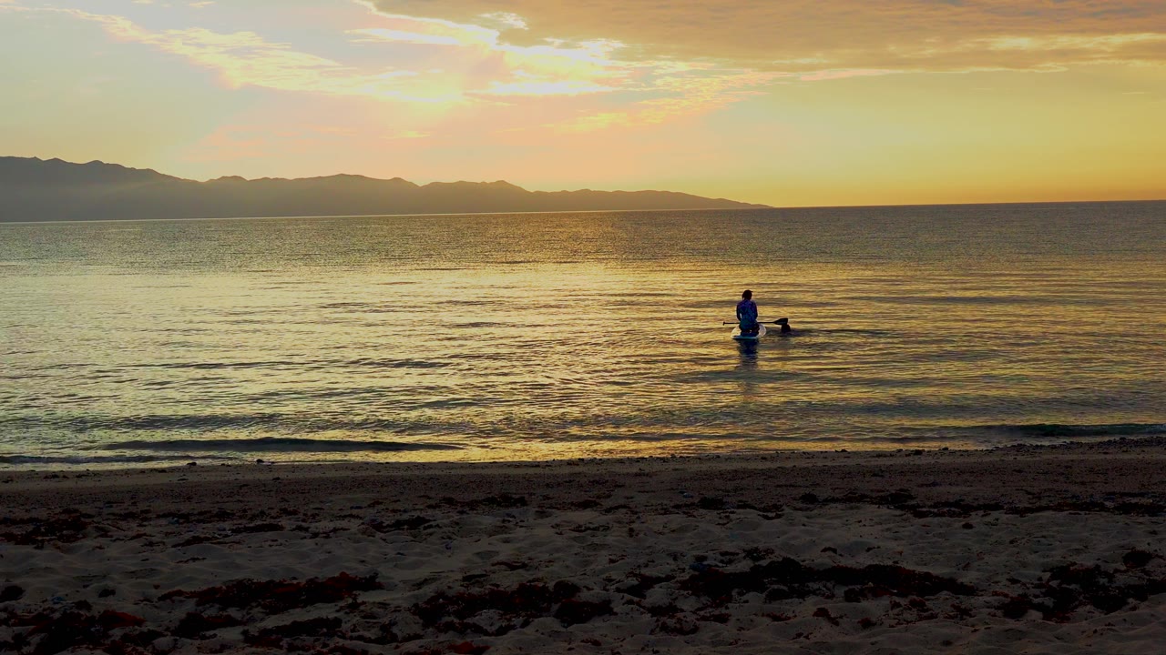 A lone paddleboarder sits farther right of center in the sunset-lit water at El Sargento La Ventana, Mexico, with gentle ripples moving toward the beach.