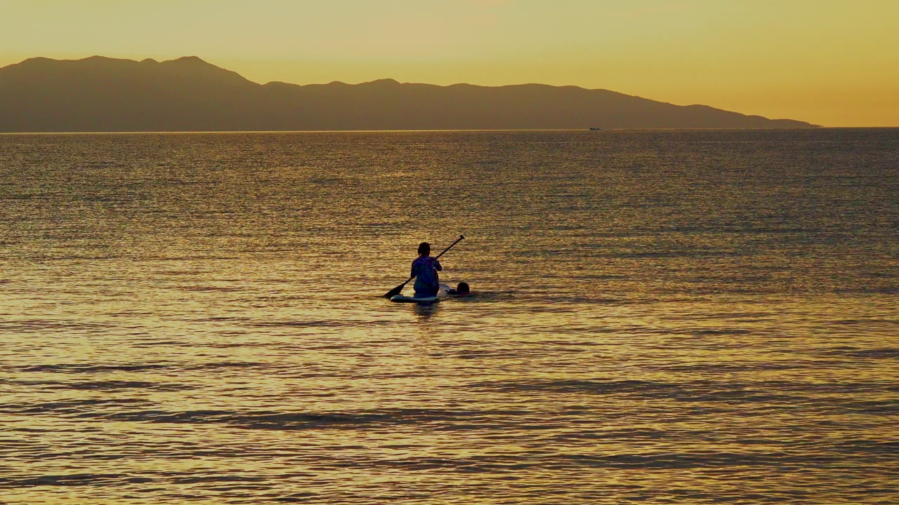 A distant paddler sits on golden water in El Sargento La Ventana, Mexico, with ripples in the foreground and dark mountain silhouettes across the horizon.