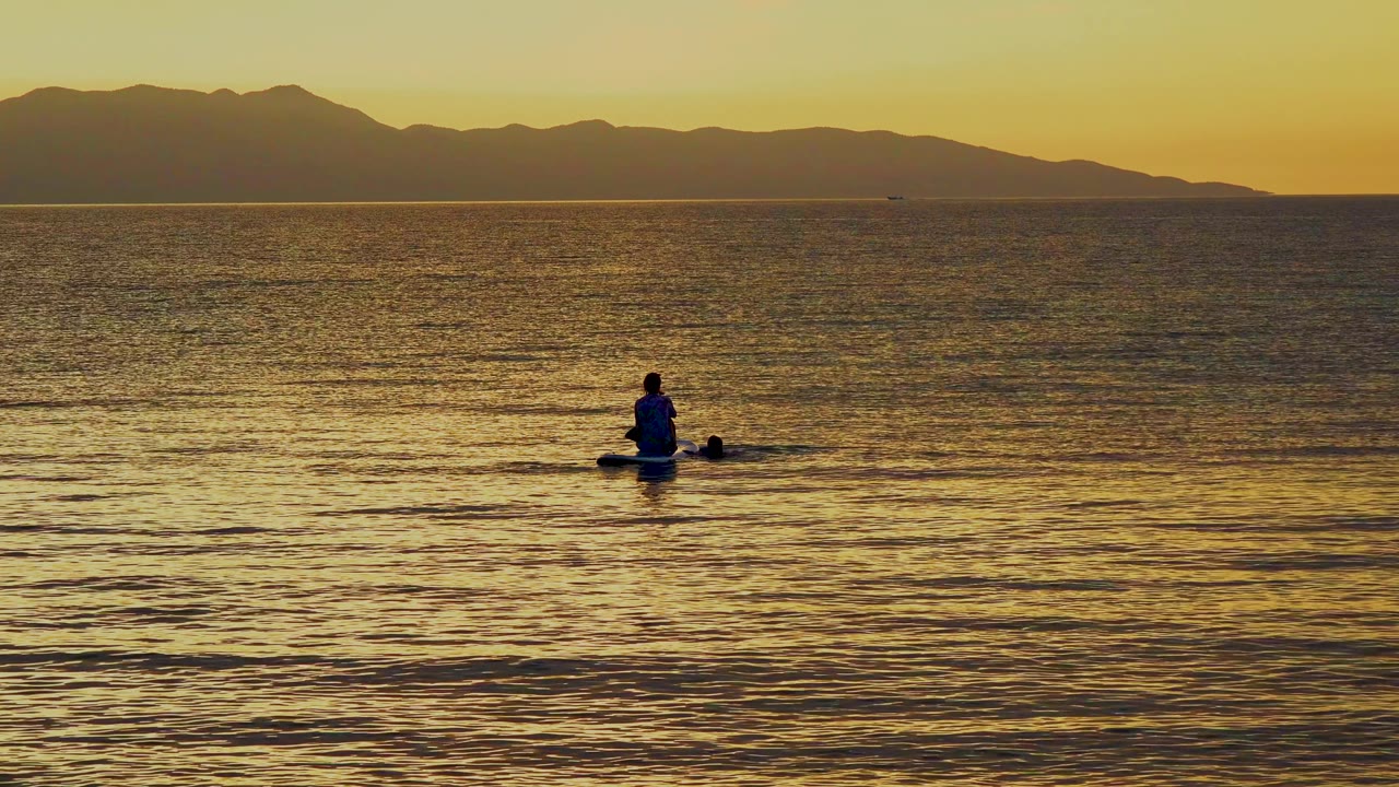 A lone distant paddler sits against golden sea in Mexico, with darker foreground ripples and a clean horizon separating the water from the mountain silhouettes.