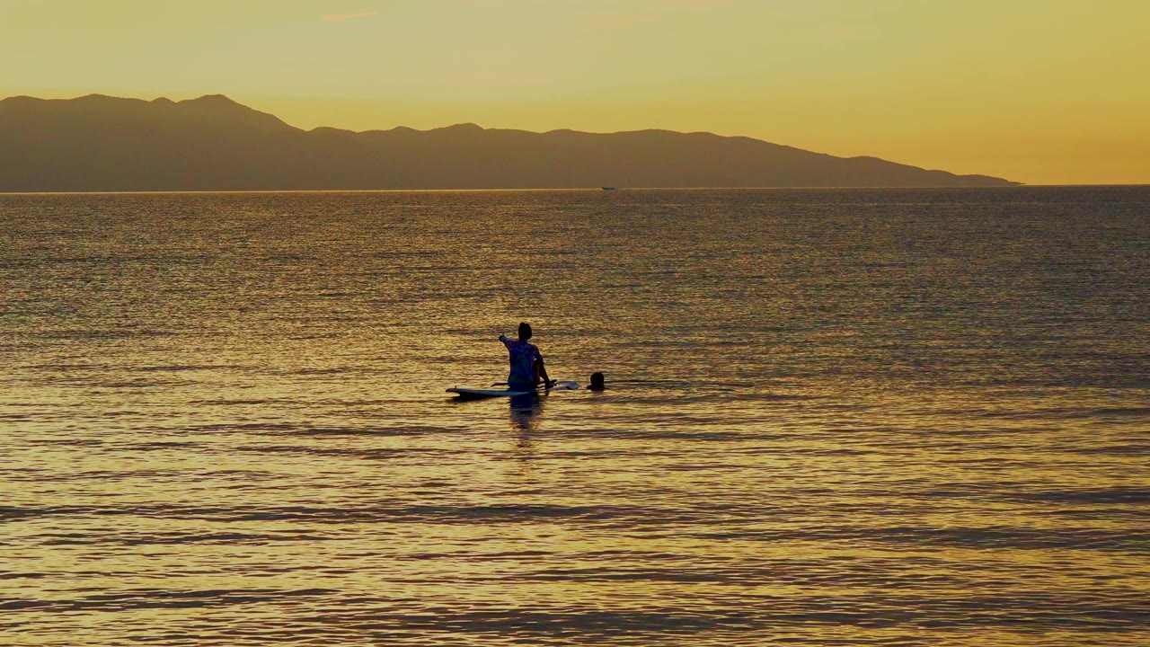 A paddle boarder sits on golden ocean water in El Sargento La Ventana, Mexico, with dark mountains across the sunset horizon and small ripples in the foreground.