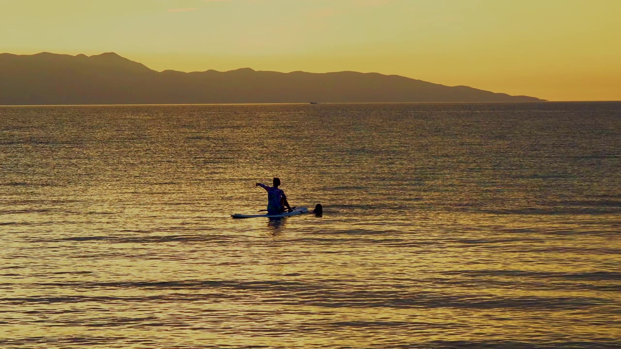 A person on a paddle board lifts an arm and paddle while drifting on golden water in El Sargento La Ventana, Mexico, with mountains silhouetted across the horizon.