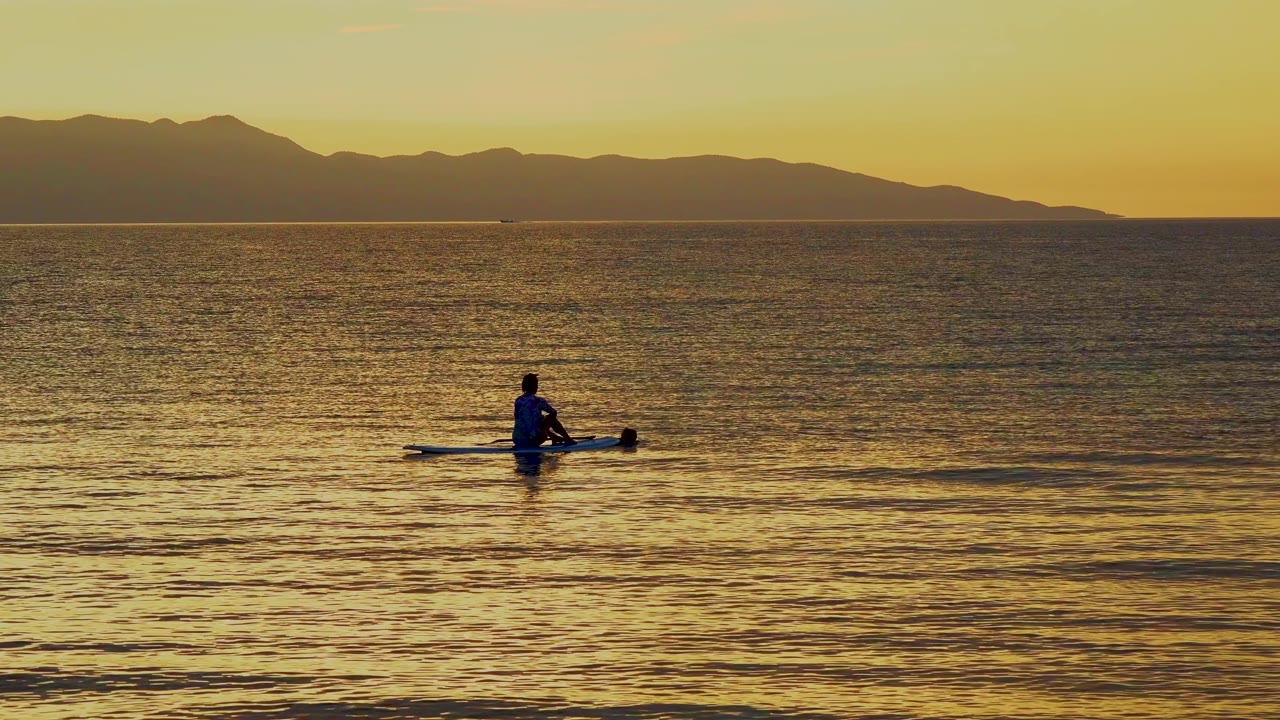 At 0:02 in El Sargento La Ventana, Mexico, the small canoeist sits on the calm sea as the amber sky brightens above the long mountain ridge.