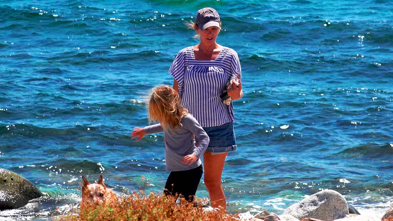 In El Sargento La Ventana, Mexico, a woman in a striped top stands by a child while a dog is partly visible near beach plants and waves roll behind them.