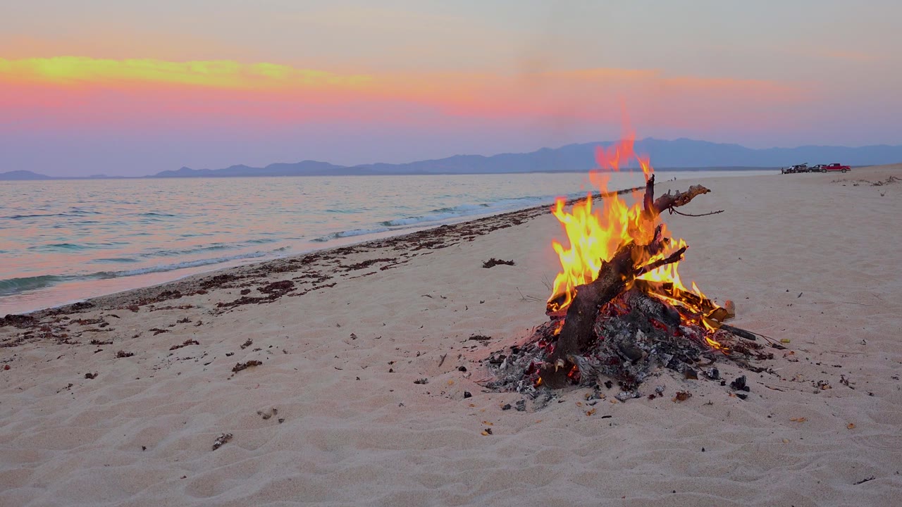 A beach campfire burns on pale sand in El Sargento La Ventana, Mexico, with calm water, distant mountains, pink sky, and no visible people.