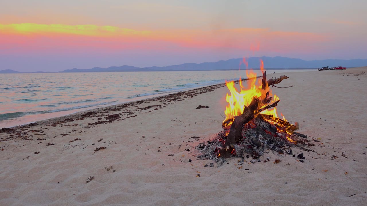Flames climb from driftwood on a sandy beach in El Sargento La Ventana, Mexico, with a diagonal waterline and no visible people.