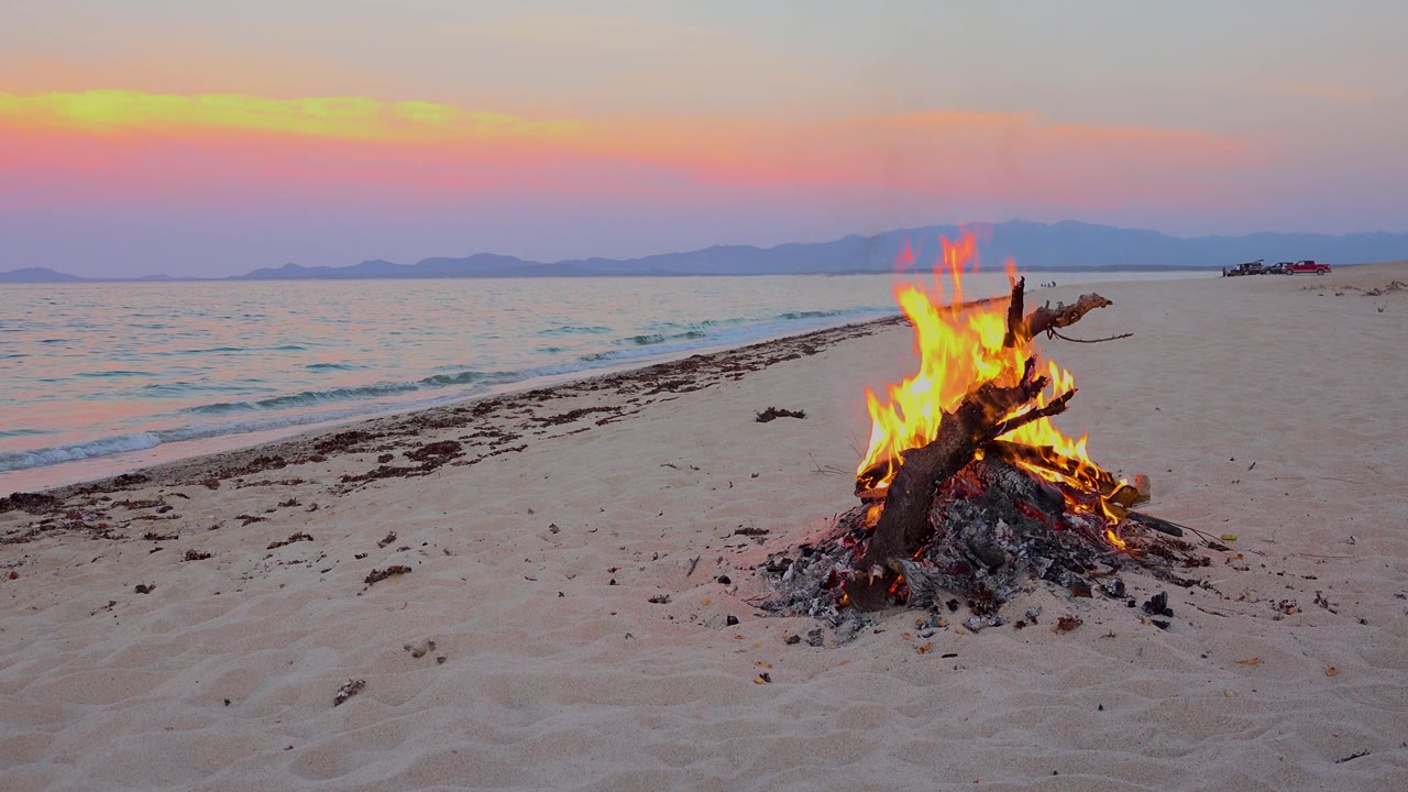 A bright campfire glows on the beach in El Sargento La Ventana, Mexico, under a pastel dusk sky with the sea at left and no visible people.