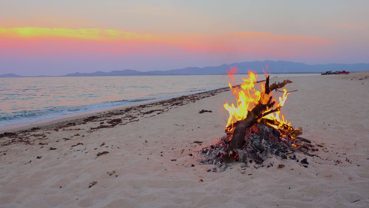 Beach campfire burning beside calm surf at sunset.