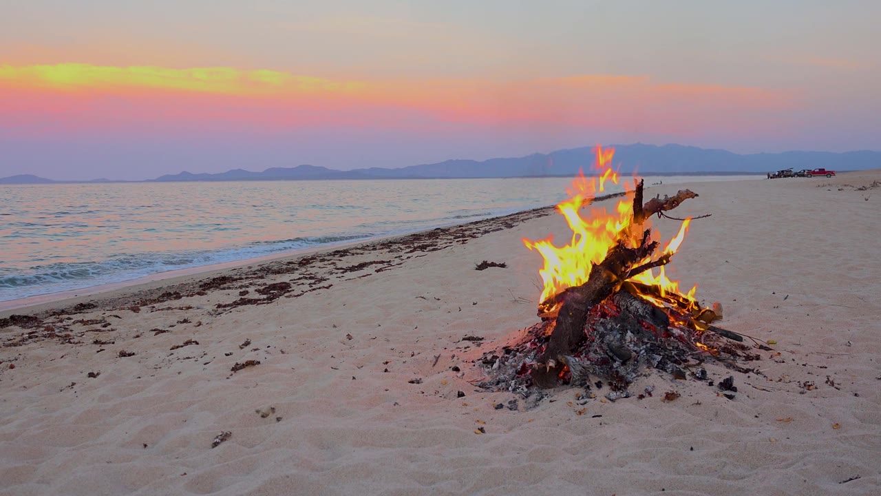 At 0:01 in El Sargento La Ventana, Mexico, flames rise from a driftwood pile on the beach while the surf sits low along the left edge and no visible people appear.