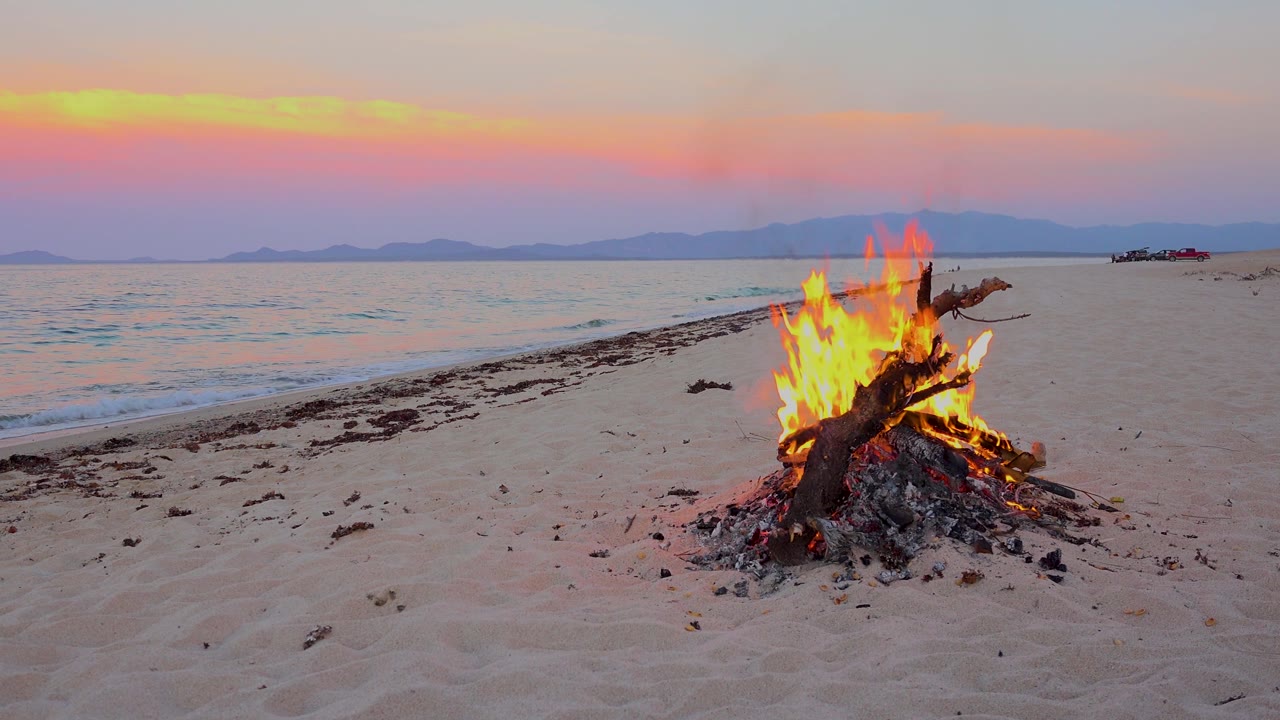 At 0:02 in El Sargento La Ventana, Mexico, the campfire flares higher against pale sand, with a thin wave breaking at left, distant mountains, and no visible people.