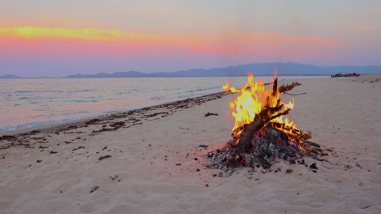At 0:03 in El Sargento La Ventana, Mexico, the fire burns lower in the foreground as a small wave curls along the shore and the empty beach continues toward distant mountains.