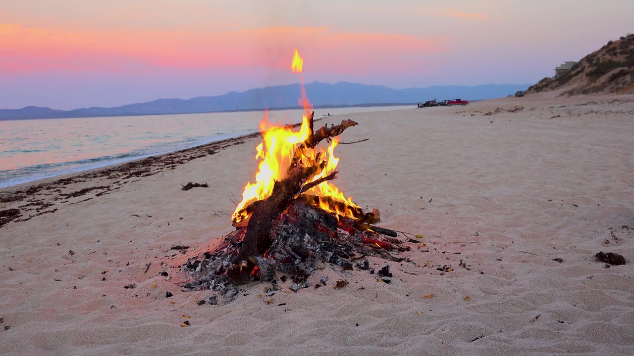 The bonfire sits slightly right of center on the sand in El Sargento La Ventana, Mexico, with a pink-purple sky, calm sea, distant mountains, and no visible people.