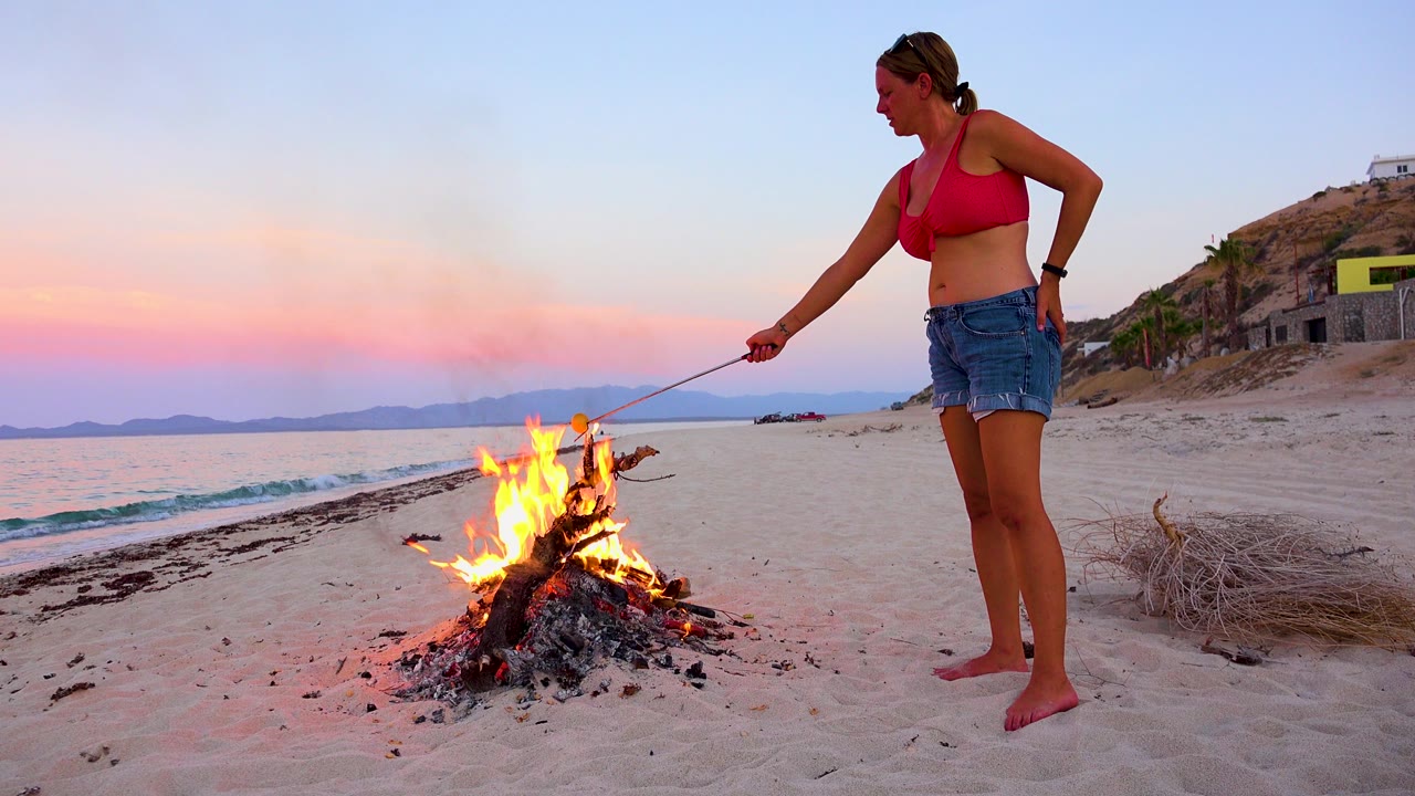In El Sargento La Ventana, Mexico, a barefoot woman in a red top stands beside a bright driftwood campfire on the sand as pink evening light colors the sky.